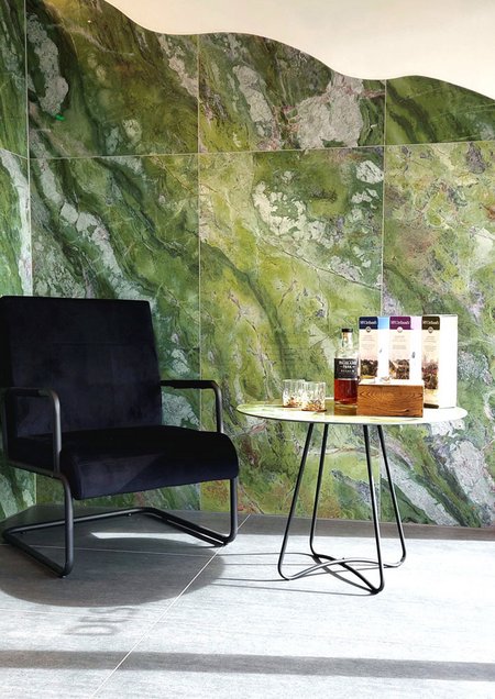 Sitzbereich mit schwarzem Sessel und kleinem Tisch vor einer markanten grünen Natursteinwand, dekoriert mit Getränken und Verpackungen.