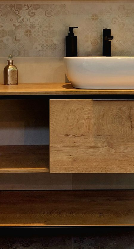 Nahaufnahme eines Waschtischs aus Holz mit weißer Aufsatzschale, schwarzen Armaturen und gemusterten Wandfliesen.