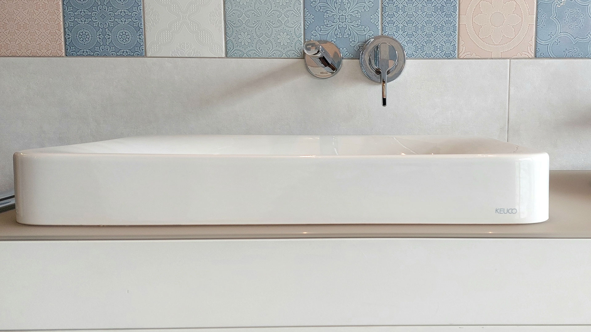 Rechteckiges, weißes Keuco-Waschbecken vor einer Wand mit farbigen, gemusterten Fliesen in Blau, Weiß und Beige. Minimalistisches Bad-Design.